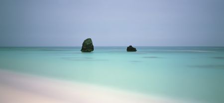 A minimalist, wide-angle seascape showing two dark rocks sitting in the middle of a perfectly still, turquoise ocean under an overcast sky.