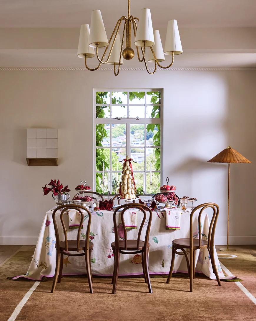 Image of a table with a white patterned table cloth with six wooden dining chairs around it in a white dining room.