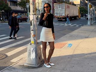 Woman poses on an NYC street wearing a white mini skirt, white The Row Mary Janes and a black fitted sweater with a crewneck t-shirt peeking out of the top.