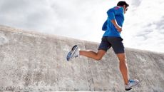 white man dark hair facing away from camera running wearing blue sleeved top dark shorts white trainers against grey wall 