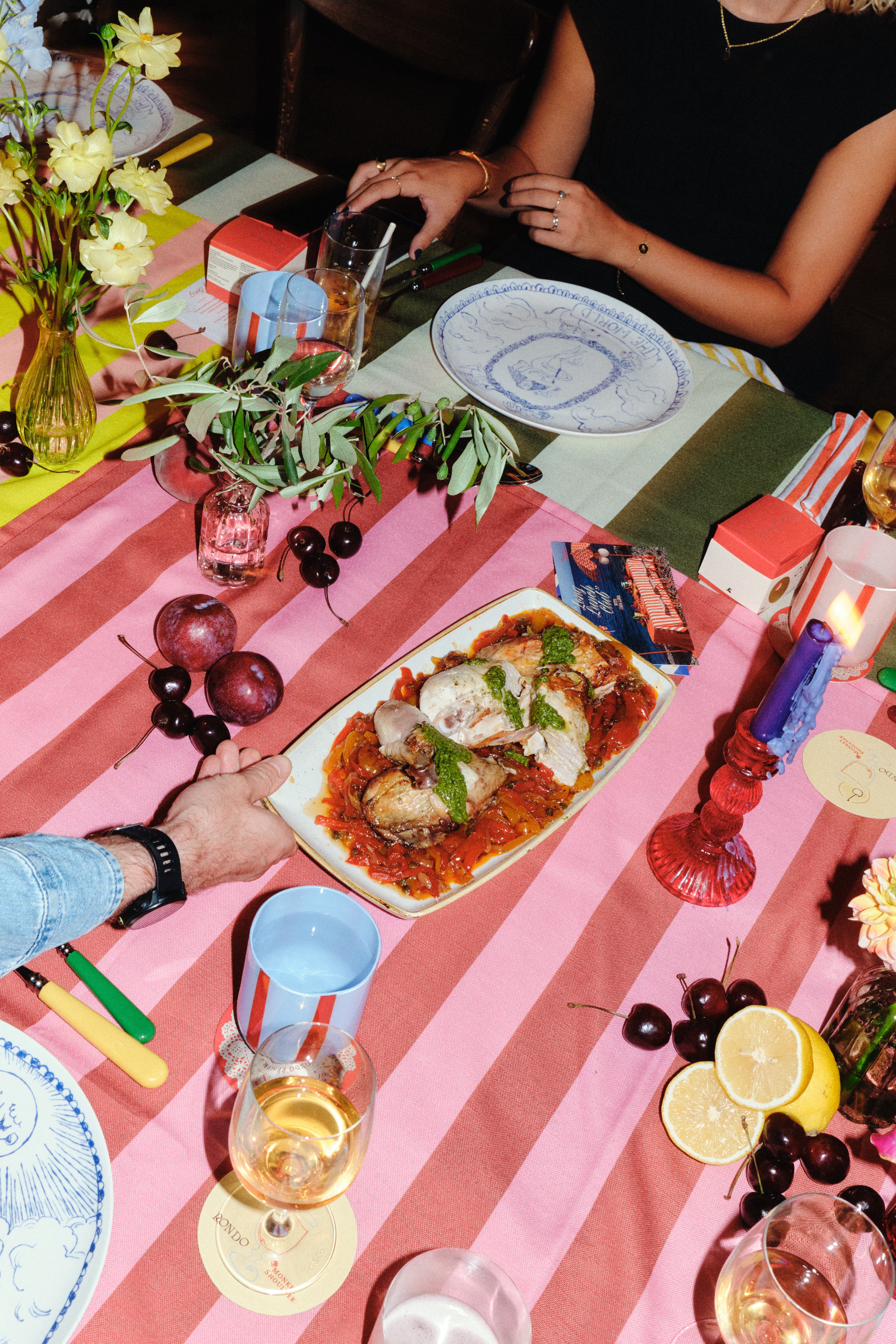 Flashed-out shot of a dinner party with silver and pastel-tinted serveware, beautifully presented dishes, striped, mismatched colorful tablecloths, and branded merchandise.