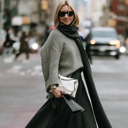 Woman crossing the street wearing sunglasses big scarf, grey sweater, white bag, and black skirt 