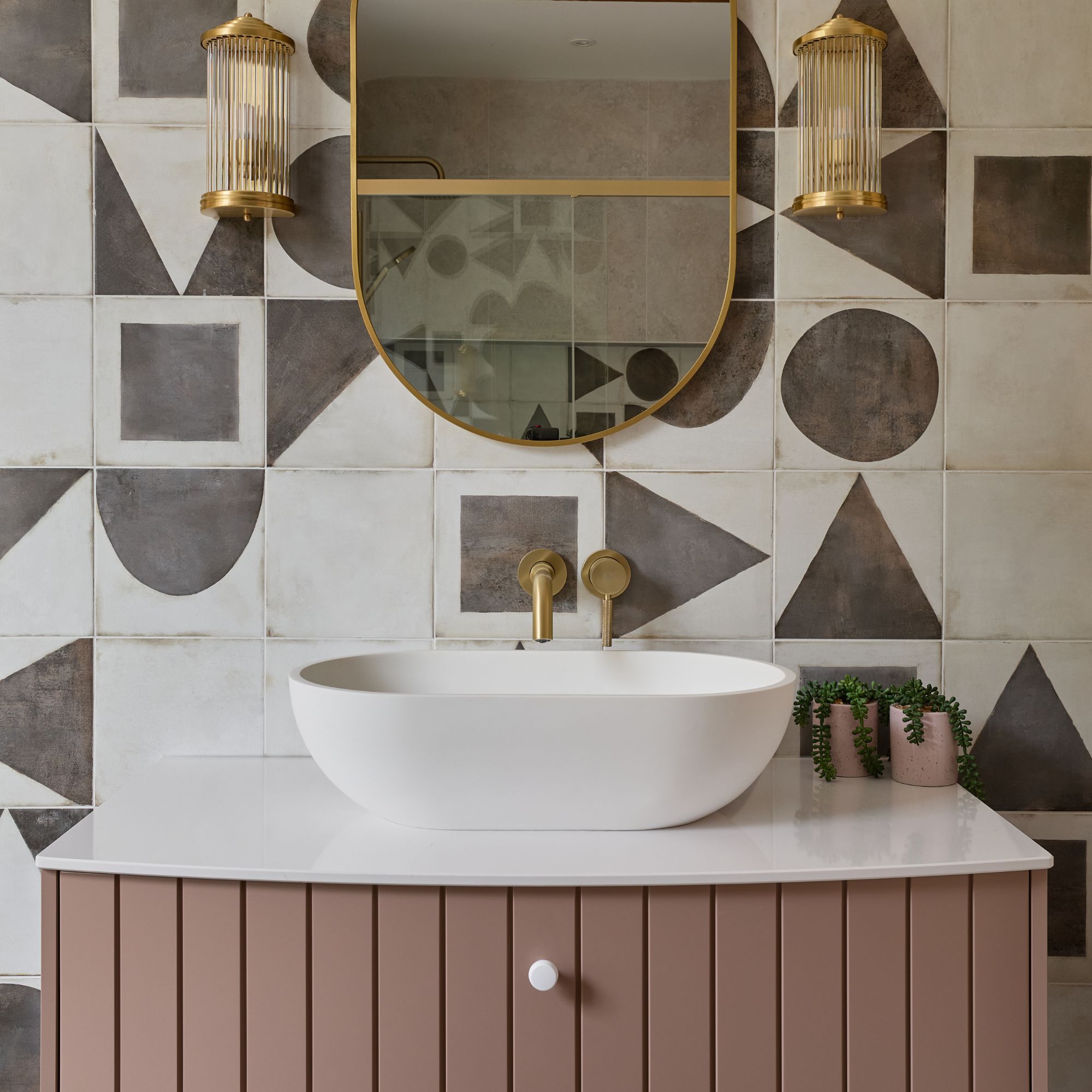 Bathroom vanity wall with gold oval mirror, wall lights, monochrome tiles and pink unit