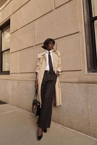 how to make a button-down shirt look more expensive is shown in a photo of a woman outside leaning against a wall wearing black glasses with a tan trench coat layered over a white button-down shirt styled with a black tie, black belt, black trousers, black kitten heels, and a black structured handbag