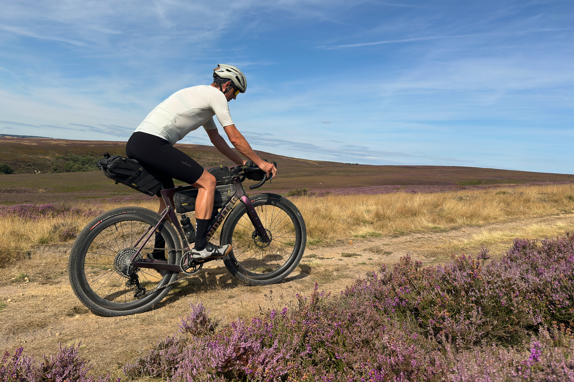 Man riding a purple gravel bike with black bikepacking bags on a sandy trail wearing grey shoes, blue shorts, a cream top and a beige helmet with heather in the foreground