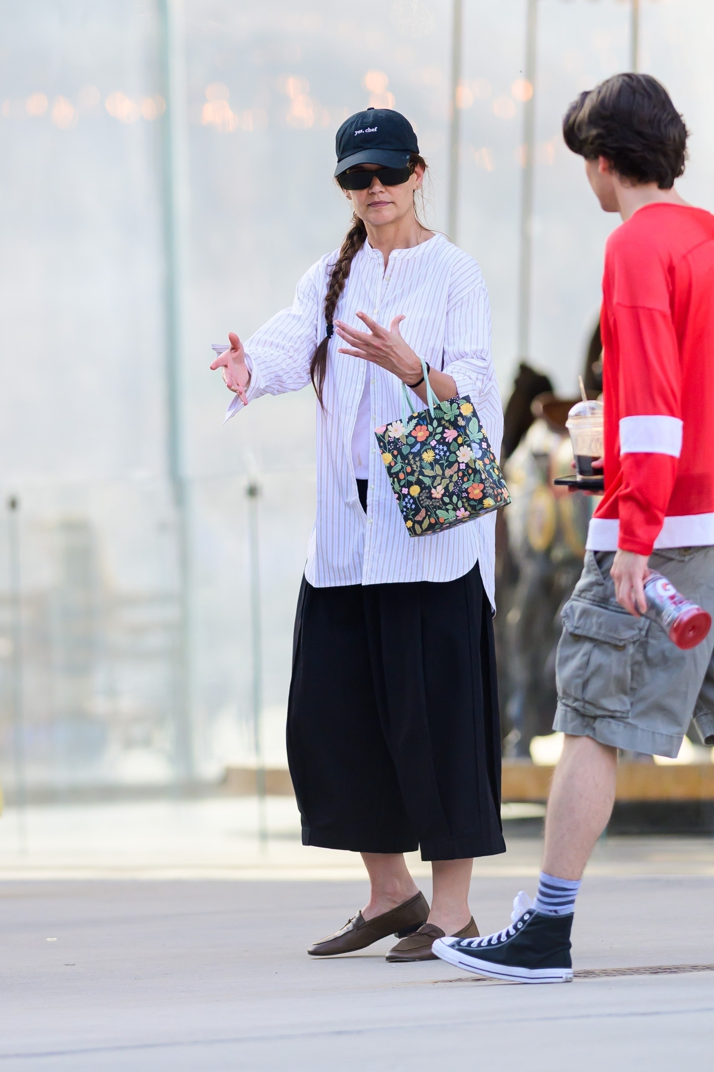 Katie Holmes wears a baseball cap, button-down shirt, black shorts, a floral bag, and brown leather loafers.