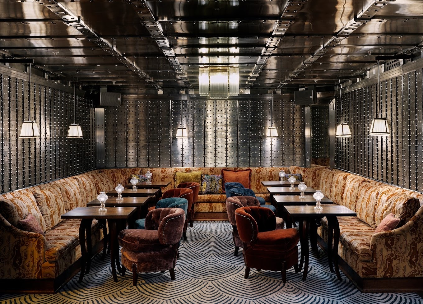 A former bank facility drenched in iron surfaces houses an intimate, characteristic cocktail bar with a U-shaped banquette seating area, velvet armchairs, Art Deco wooden tables and lighting, and a central glass chandelier.