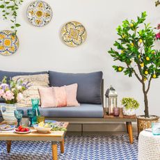 Patio with metal and wooden L-shaped sofa on a blue and white outdoor rug with coffee table. Faux lemon and cherry trees and plates on the white wall