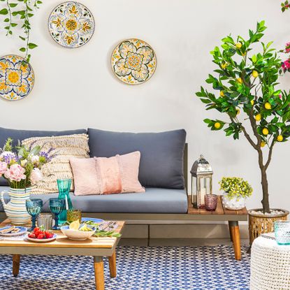 Patio with metal and wooden L-shaped sofa on a blue and white outdoor rug with coffee table. Faux lemon and cherry trees and plates on the white wall