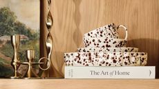 A stack of splatterware dinnerware on a homeware book displayed on a wooden shelf next to two gold candlesticks