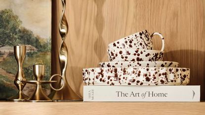 A stack of splatterware dinnerware on a homeware book displayed on a wooden shelf next to two gold candlesticks