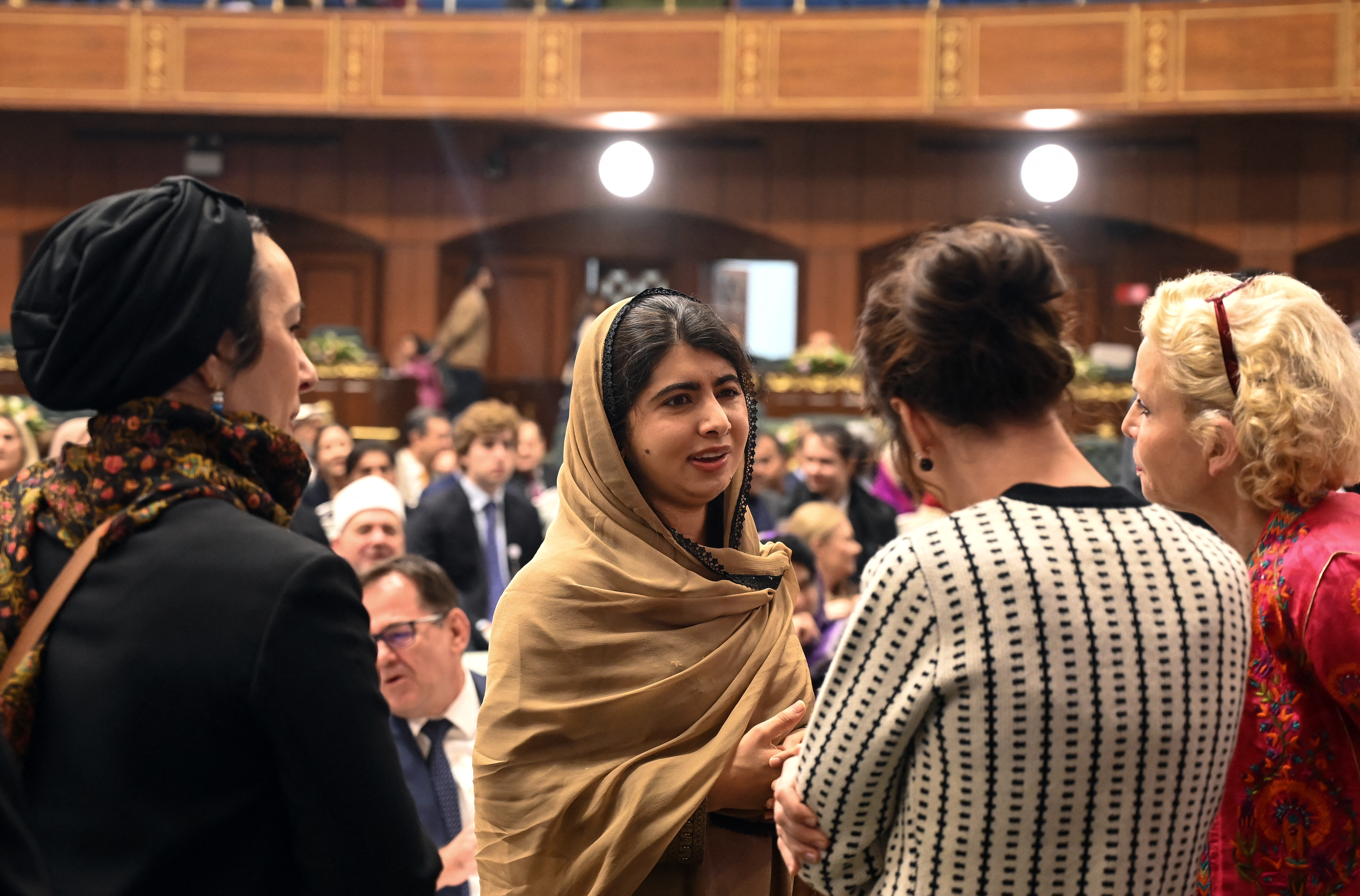 Malala Yousafzai speaks with attendees at a formal event, wearing a tan headscarf and surrounded by guests in conversation.
