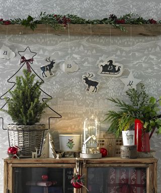 country christmas decorations on a wooden display cabinet in front of a wall with wallpaper featuring alpine scenery, mini fir trees and carved wood houses and wildlife, underneath a wooden beam with foliage and berries garland