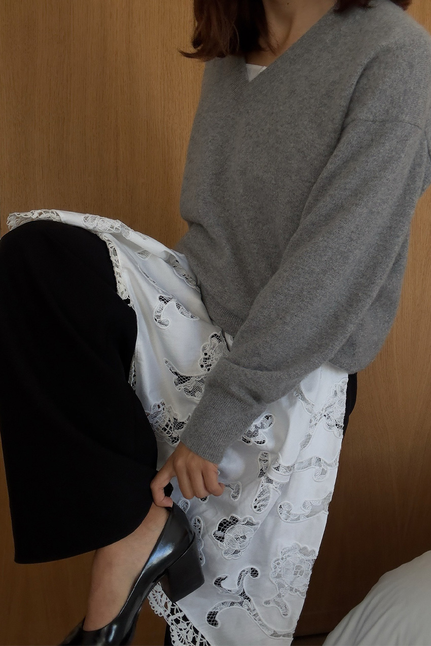 A sweater styling trend is shown in a close photo of a woman&#039;s outfit, where she&#039;s standing in a hotel room wearing a gray sweater over a white lace dress worn over black jeans styled with black kitten heel pumps
