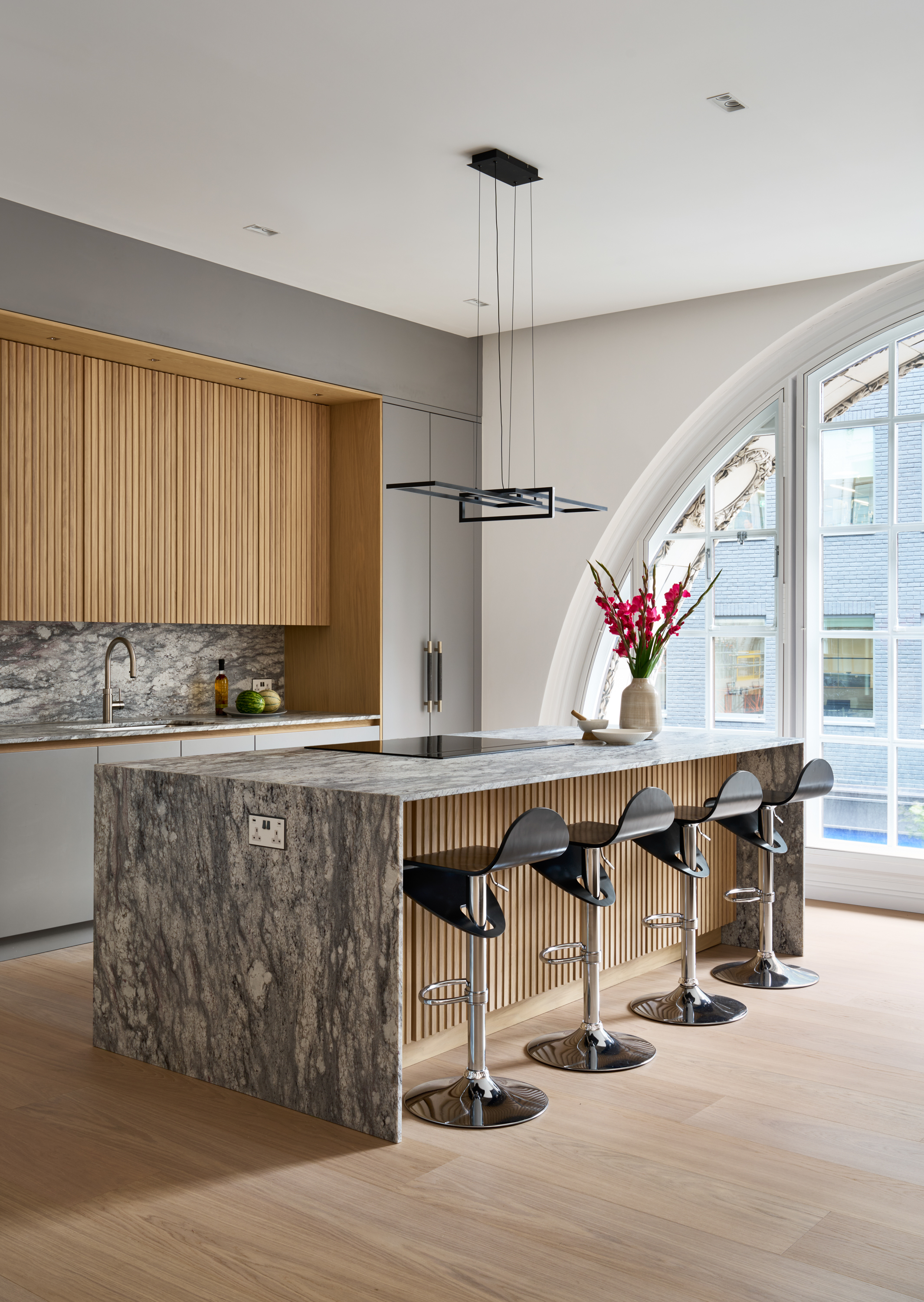 A modern kitchen with medium gray ceiling height kitchen cabinets with a fluted wood alcove and marble backsplash and kitchen island that has the same fluted wood detailing with four black bar stools in front and a vase of flowers on top