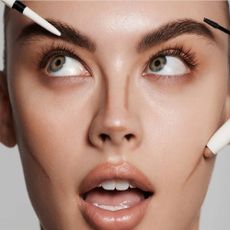 A close-up of a model's face with makeup brushes and pens around the face.