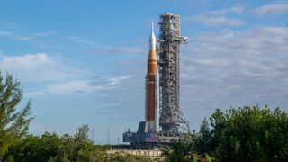 An orange rocket held up by scaffolding sits behind large green bushes under a blue sky.
