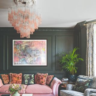 A dark green-painted living room with wall panelling, a pink velvet sofa and a sculptural pink chandelier