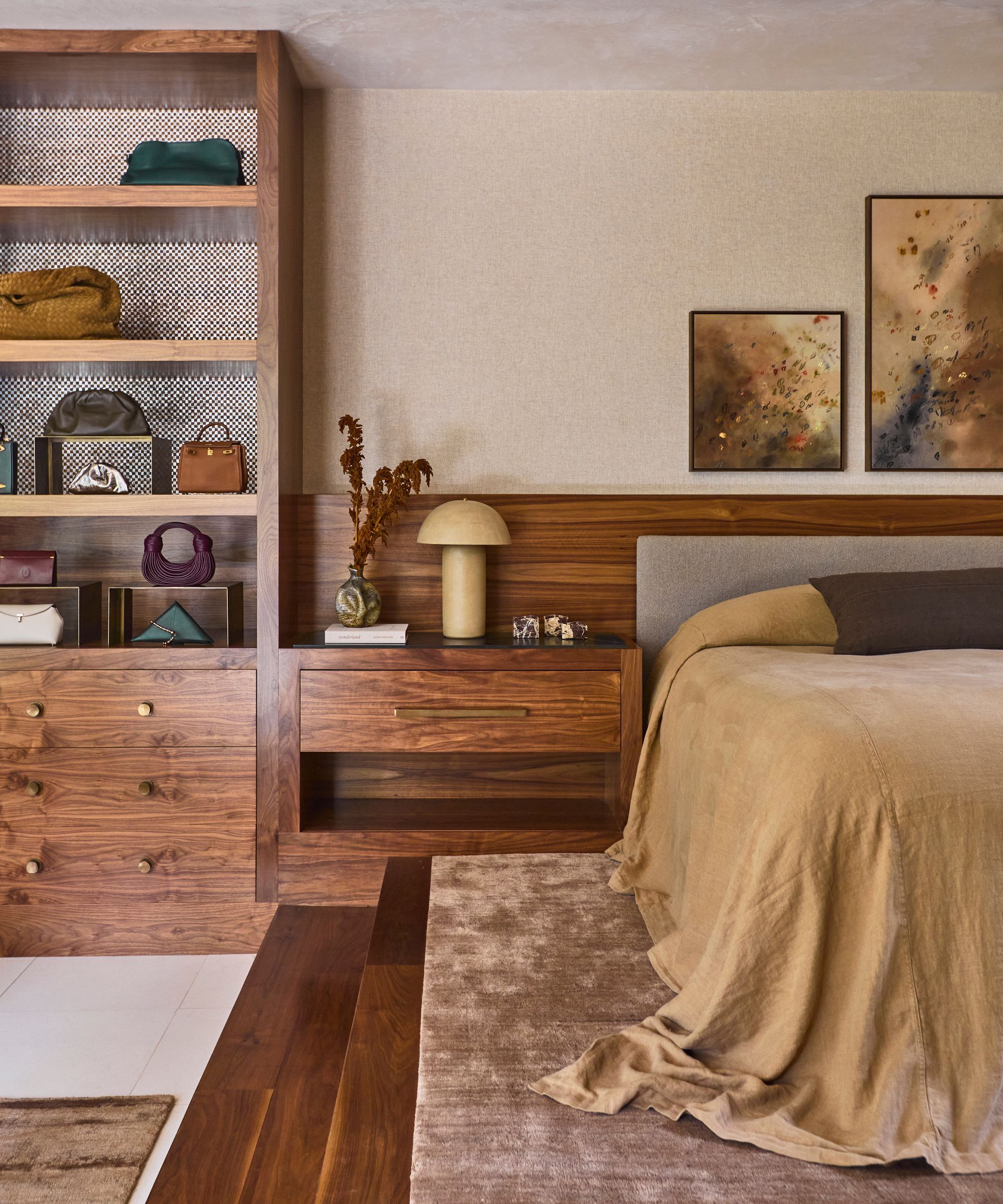 modern bedroom with raised platform, timber stairs, timber built-in headboard and joinery with handbags on display, a mushroom lamp and vase of flowers on side table, artwork above bed, and bed made with oversized linen bedcover