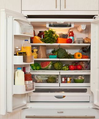 An open fridge with fruit and vegetables on the shelves and milk and juice bottles in the door, with cream cabinets around it