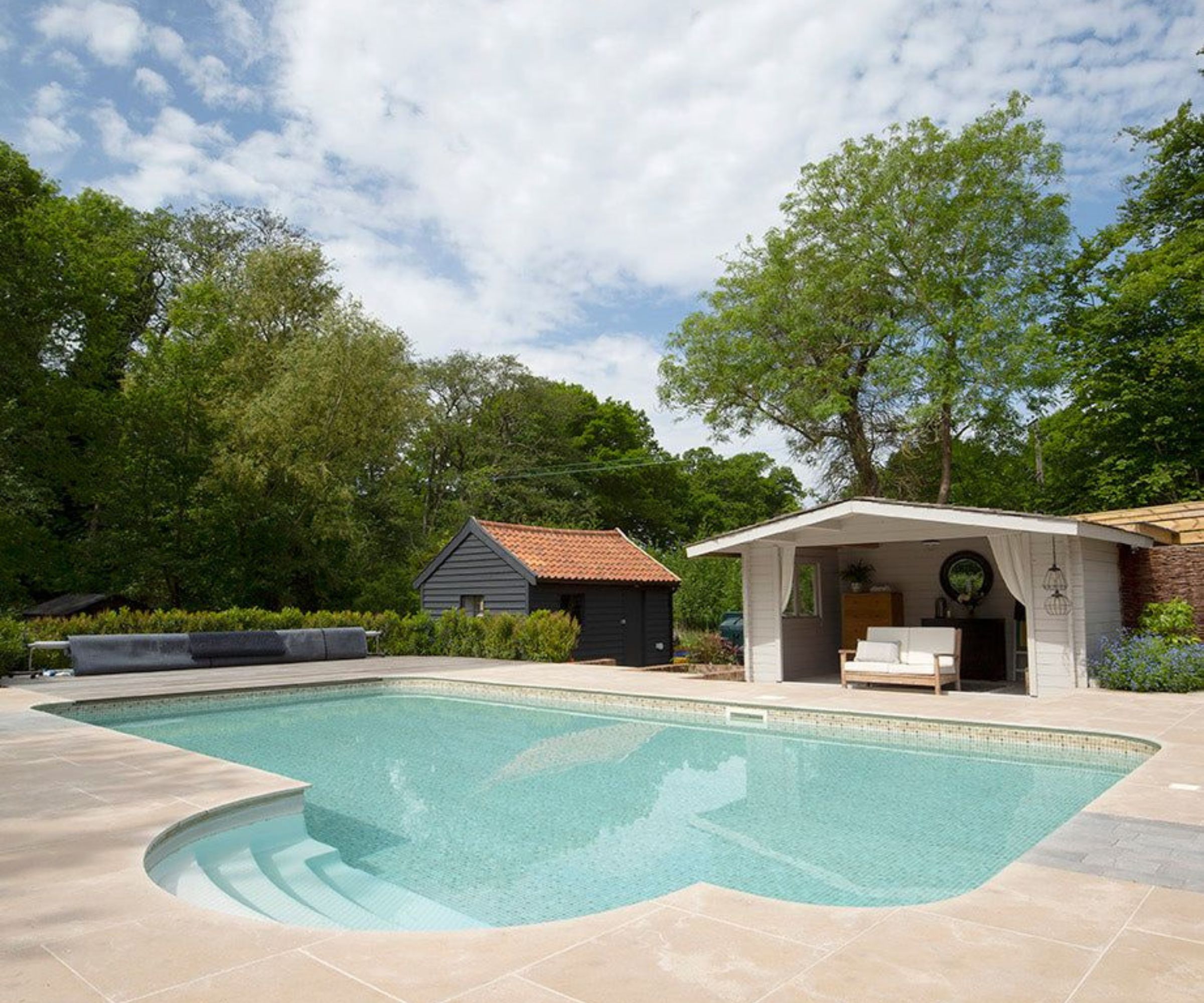 A freeform outdoor swimming pool with limestone paving, a white open-fronted pool house with teak sofa, a dark barn beyond and woodland backdrop.