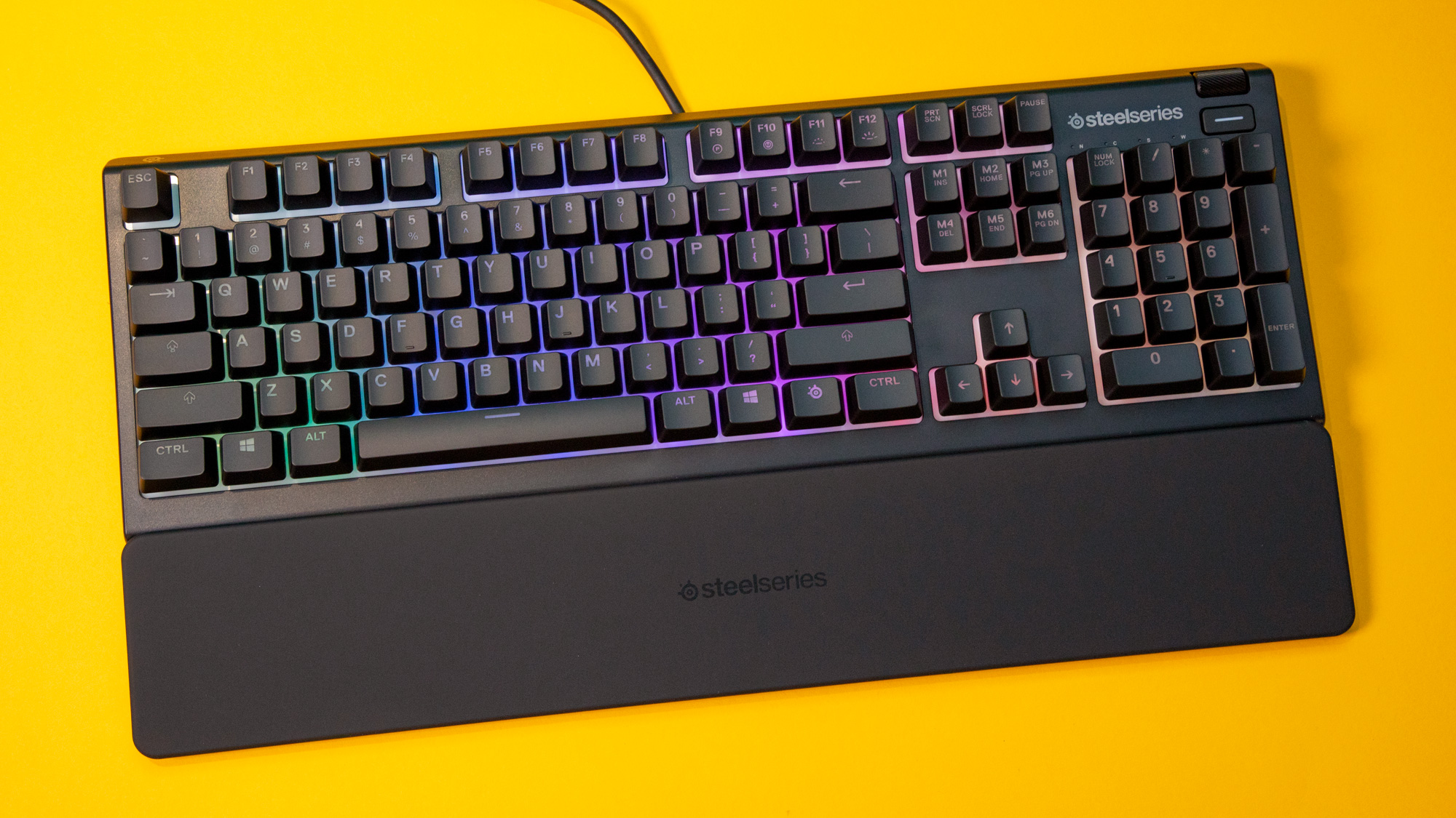 Et sort SteelSeries Apex 3-tangentbord ligger på ett gult bord.