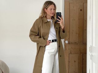 British influencer Alexis Foreman poses for a mirror selfie in a bright bedroom wearing a classic tan trench coat, white t-shirt, black belt, and white jeans