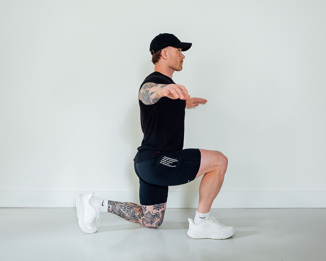 Coach Corey Perkins performs a half-kneeling pose with an upright torso. His left leg is bent at a 90-degree angle, with his toes and knee resting on the floor. His right leg is also bent at a 90-degree angle, but his right knee is elevated in line with his hip and the sole of his right foot is planted on the floor. He holds his chest up and proud and his arms out to the sides.