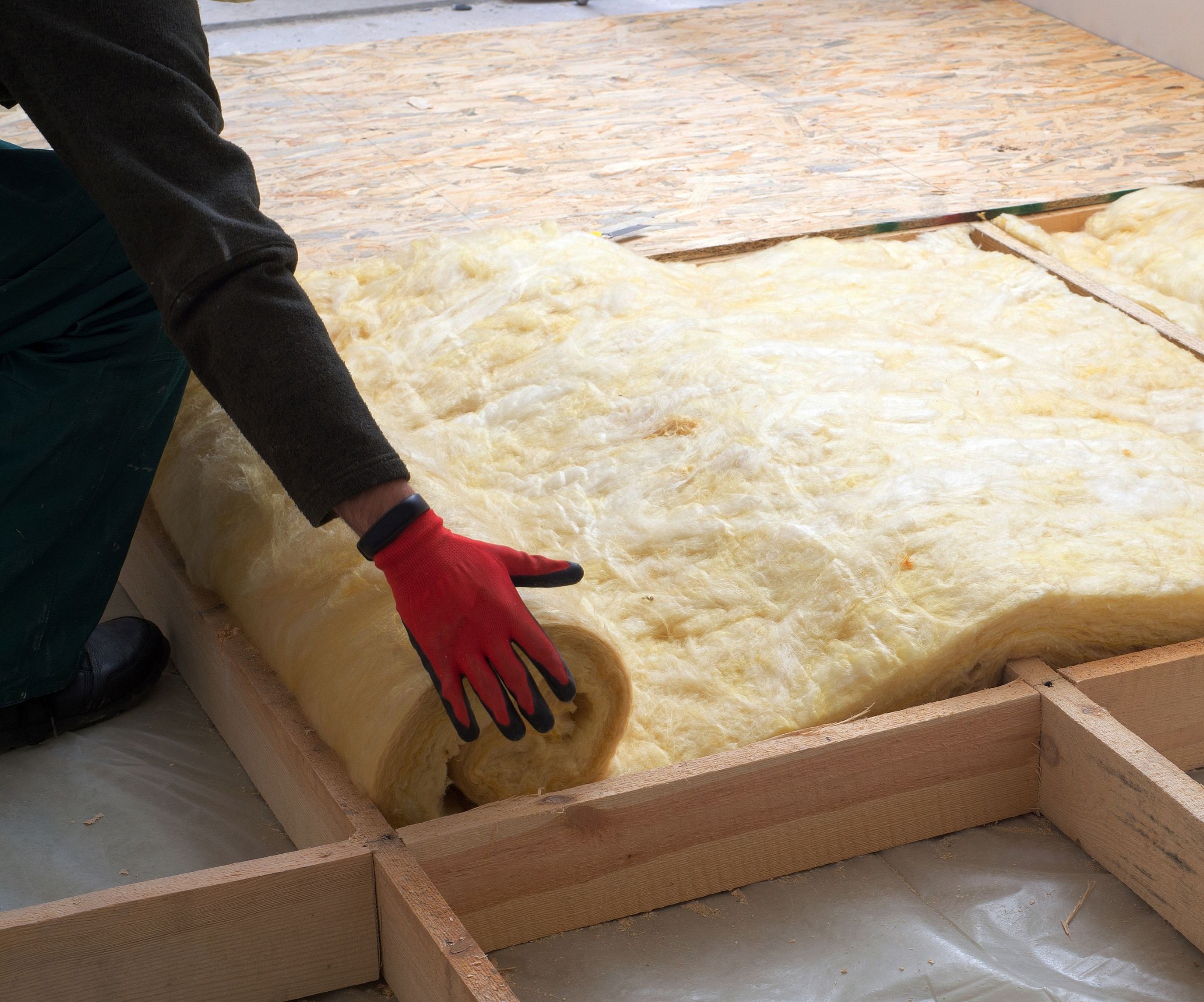 Insulation being rolled out by a man in a dark jumpsuit and red gloves over flooring joists