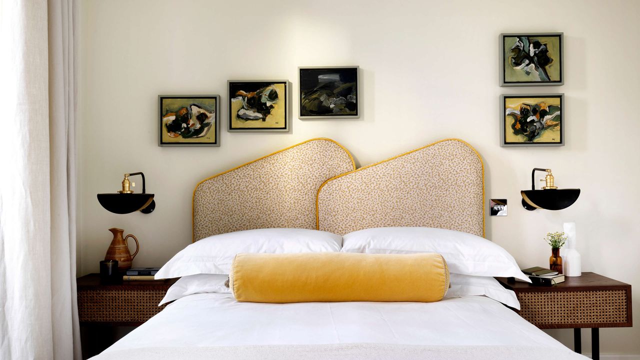 Pandora Taylor yellow shapely headboard with white bed sheets, one of the bed sheet colors to avoid, with artwork on the walls and yellow bolster cushion