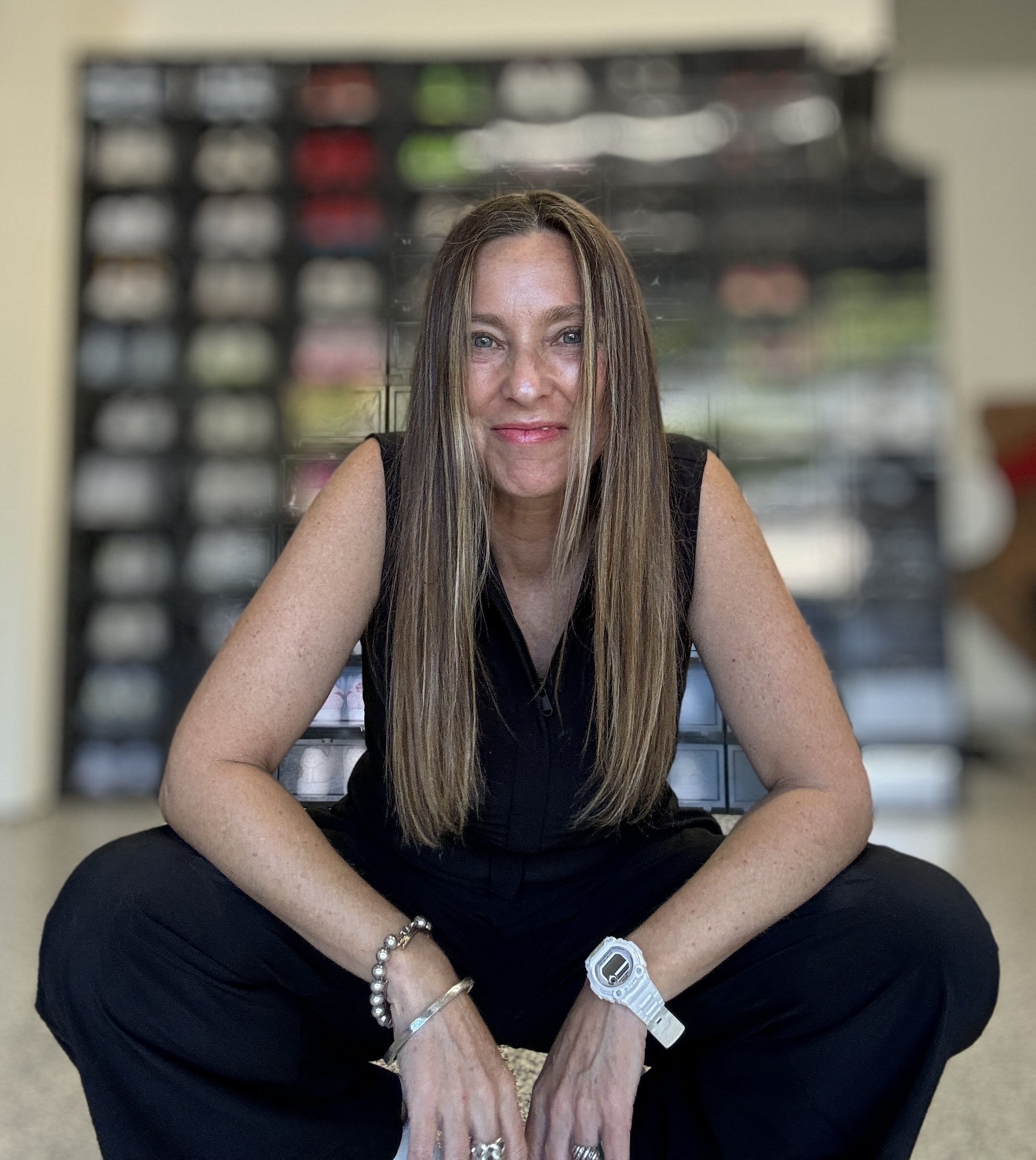 professional organizer Michelle Urban sat on the floor in a cream colored room in front of a black shelf filled with yarn: a lady with long mouse brown hair wearing all black smiling at the camera sitting on the floor