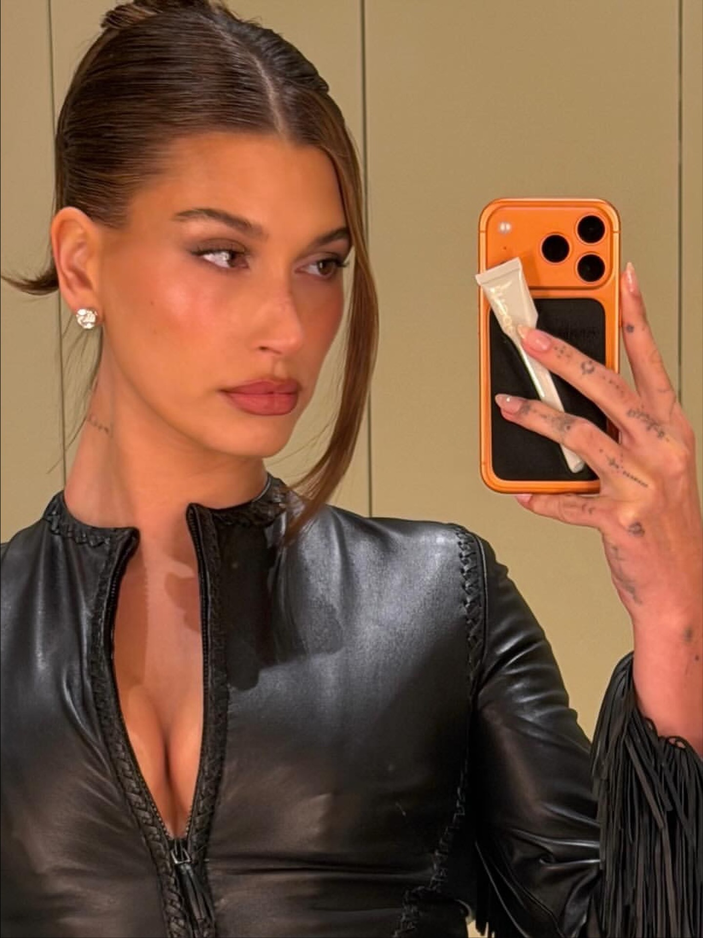 Hailey Bieber in a leather top taking a selfie in a mirror with peach French tip nails