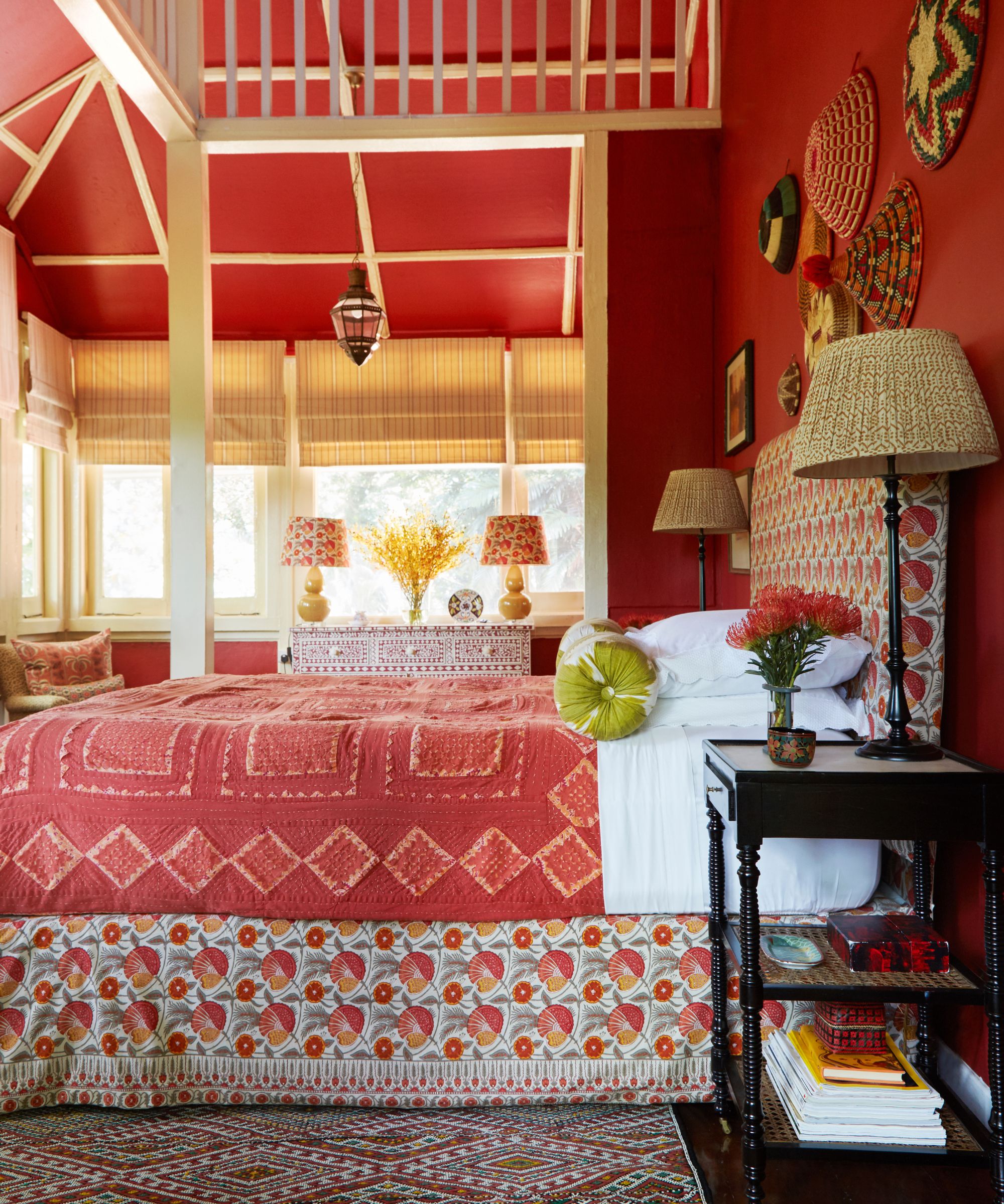 A side-on view of a bright red bedroom, with a large bed with floral-patterned bed with white and red bedding, two matching lamps on either side, a black bedside table seen in the foreground with a stack of books, vase of red flowers, and other miscellaneous items seen, beside a large patterned rug. In the background, bright windows are seen, with a patterned chest of drawers, yellow flowers, and two yellow lamps with patterned shades on top, and above a mezzanine is seen.