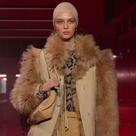 A model wearing a taupe cap, brown fur coat, and patterned blouse walking at the F/W 25 Valentino show during Paris Fashion Week.