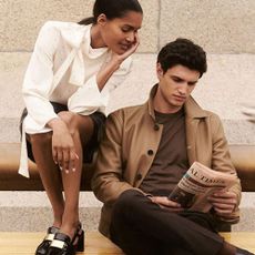 A man sat on a bench reading a newspaper with a woman perched on the back of the bench, peering over his shoulder at the newspaper, both wearing clothes from Reiss