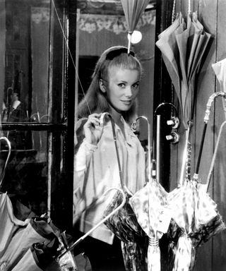 catherine deneuve looking through a store window with umbrellas in it in The umbrellas of Cherbourg