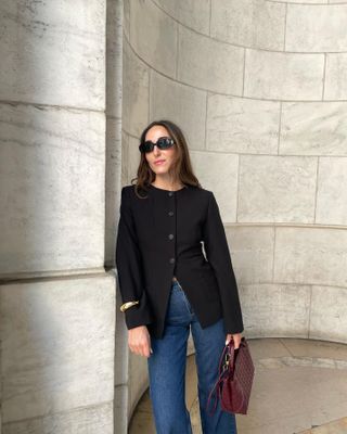 Woman wearing black collarless jacket, jeans, black sunglasses and red bag standing in front of marble hallway.