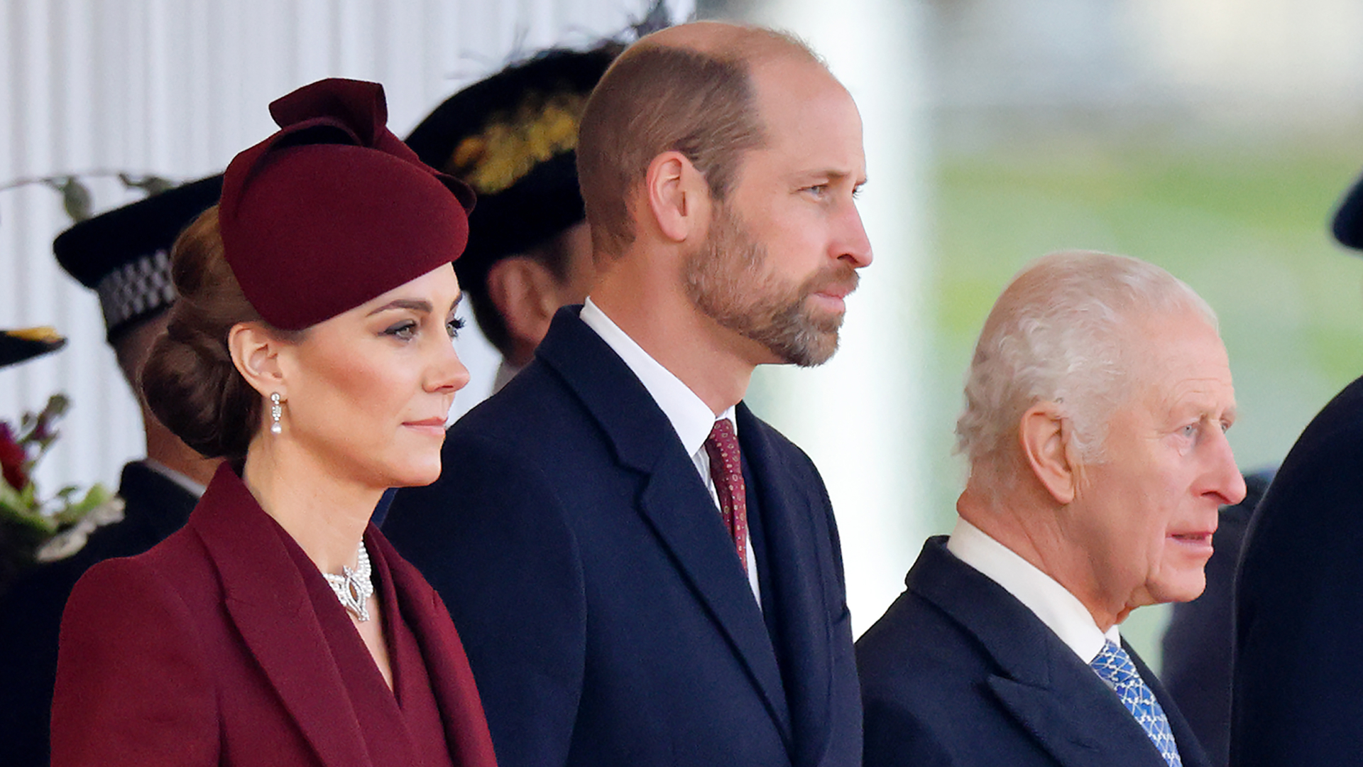 Kate Middleton wears a burgundy coat and matching hat, Prince William wears a navy coat and red tie, and King Charles wears a navy coat with a white shirt and blue tie