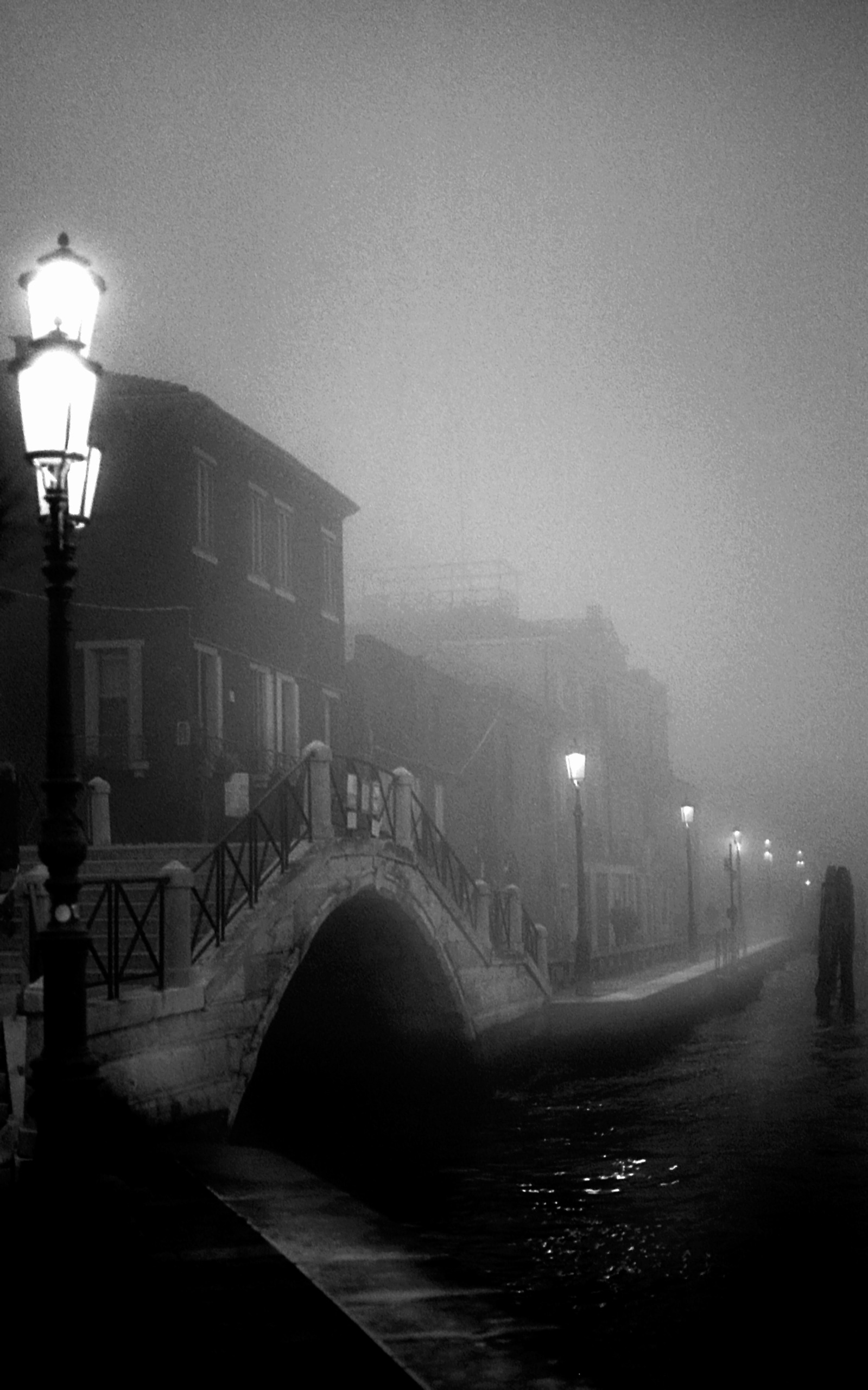 An arched stone bridge crosses a Venetian canal at night, with a row of street lamps fading into thick fog.