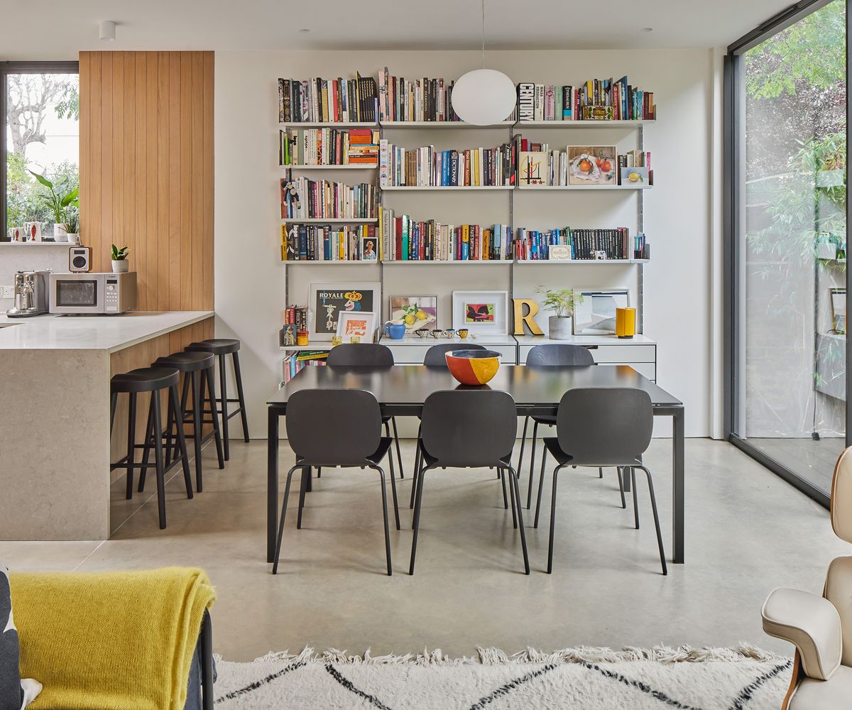 A black dining table next to a kitchen island with a wall of shelving holding lots of bright colourful books and small framed art pieces