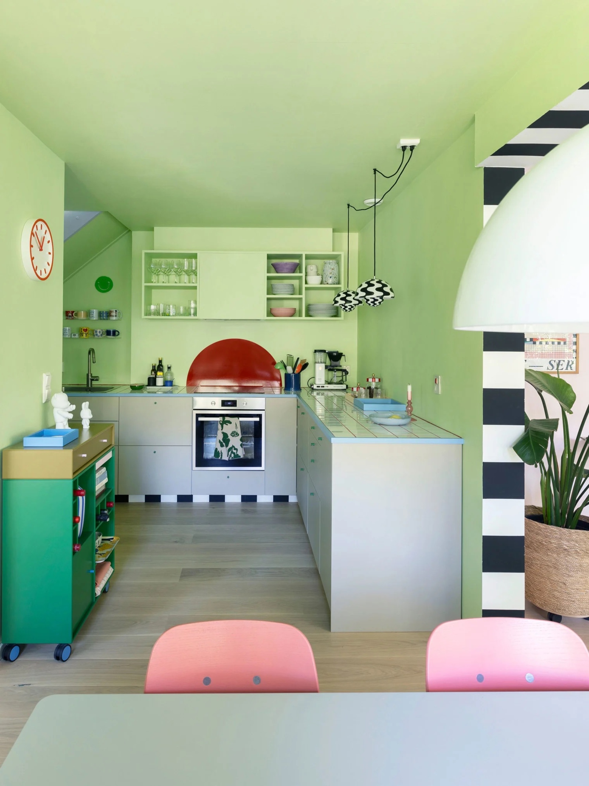colorful kitchen color drenched with light sage green, gray cabinetry with blue counter, open shelving, black and white zig zag pendant lights, portable green storage, wall clock and dining table in foreground with pink chairs
