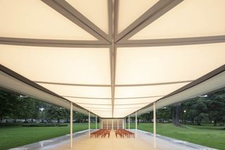 Interior view of the white framed pavilion, white floor, red metal benches and surrounding view of the lawn and trees
