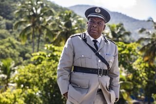 Commissioner Selwyn Patterson (Don Warrington) standing in his uniform with a canopy of green trees in the background, with a concerned look on his face