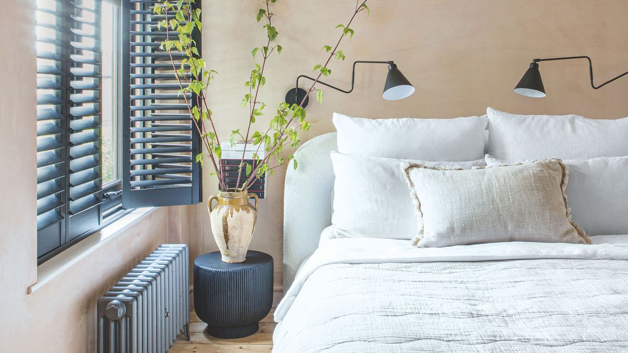 Neutral bedroom with plain plaster walls, white bedding, black radiators and shutters and accessories