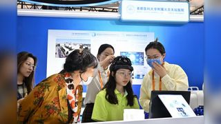 a woman wearing a polo sits between four other women at a trade fair as they adjust a BCI system on her head