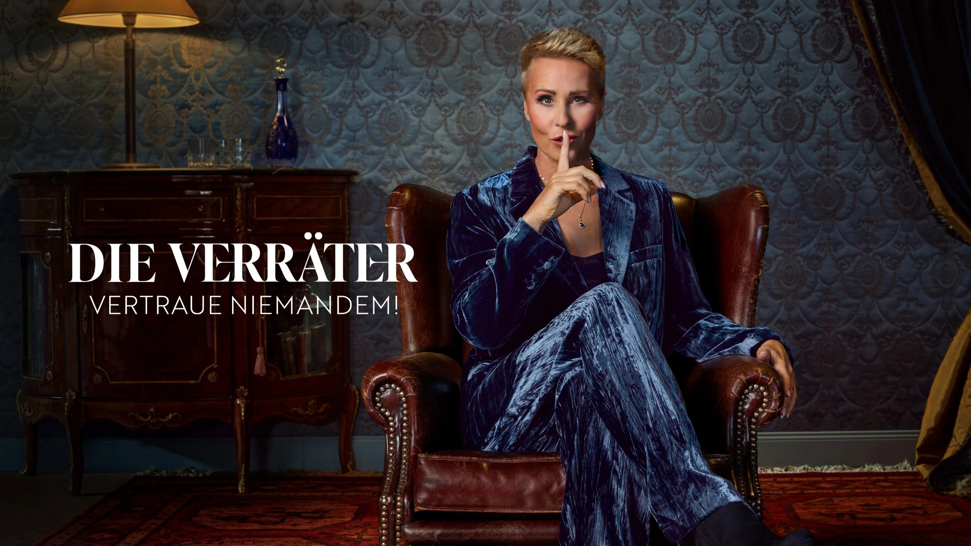 A person with short blonde hair sits in a leather armchair and holds a finger to their lips in a "shush" gesture, positioned in a dimly lit room next to the German text "Die Verr&amp;auml;ter &amp;ndash; Vertraue Niemandem!"