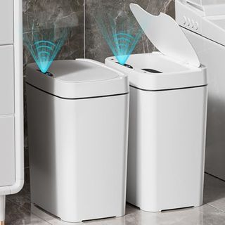 Two white plastic trash bins with automatic lids, one open one closed, in a bathroom 