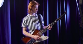 Toby Lee stands in front of a blue velvet curtain and wears a Breton stripe T-shirt and plays a Gibson Firebird.