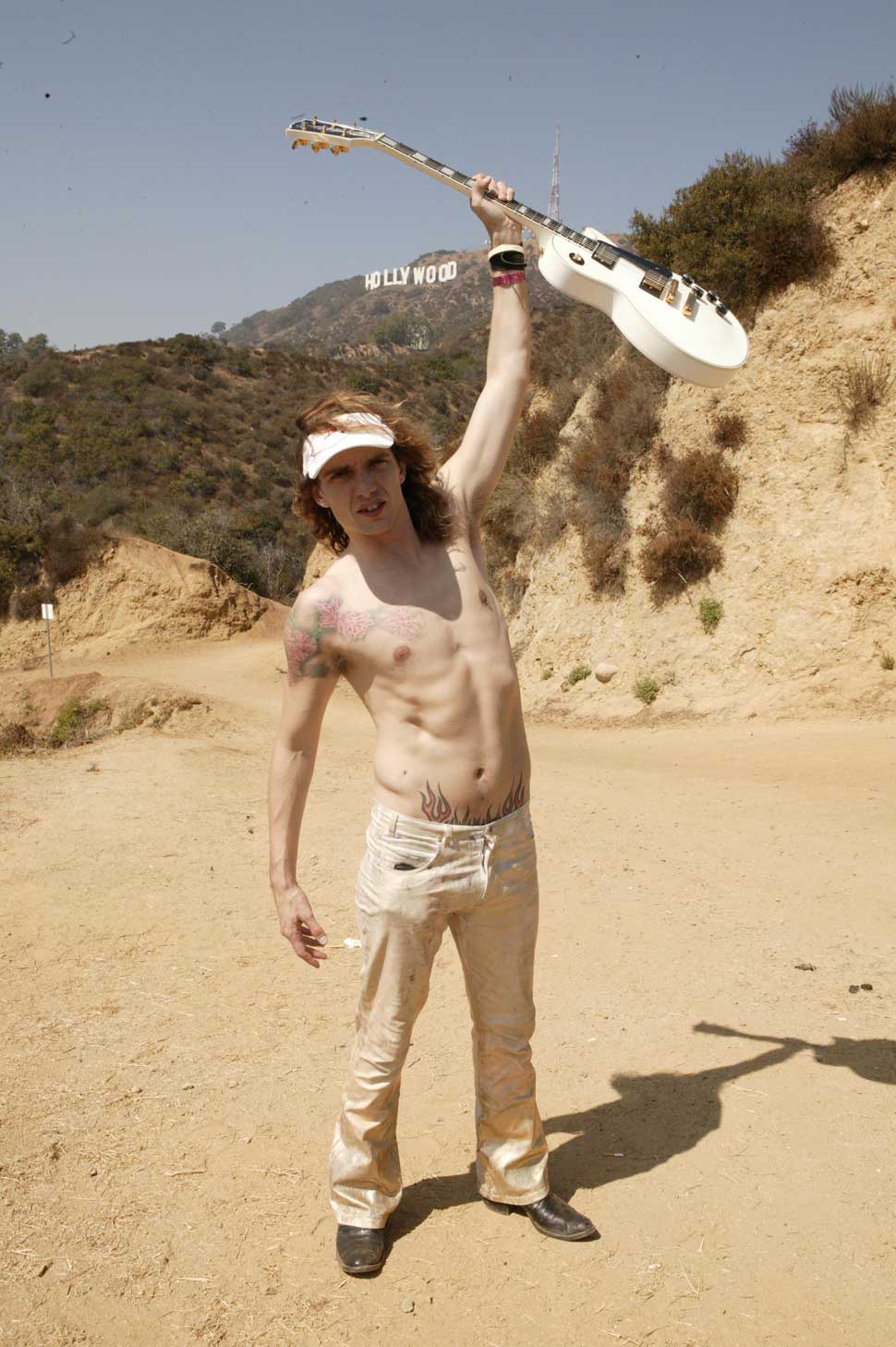 A topless Justin Hawkins brandishing a guitar on a beach in Los Angeles with the Hollywood sign in the background