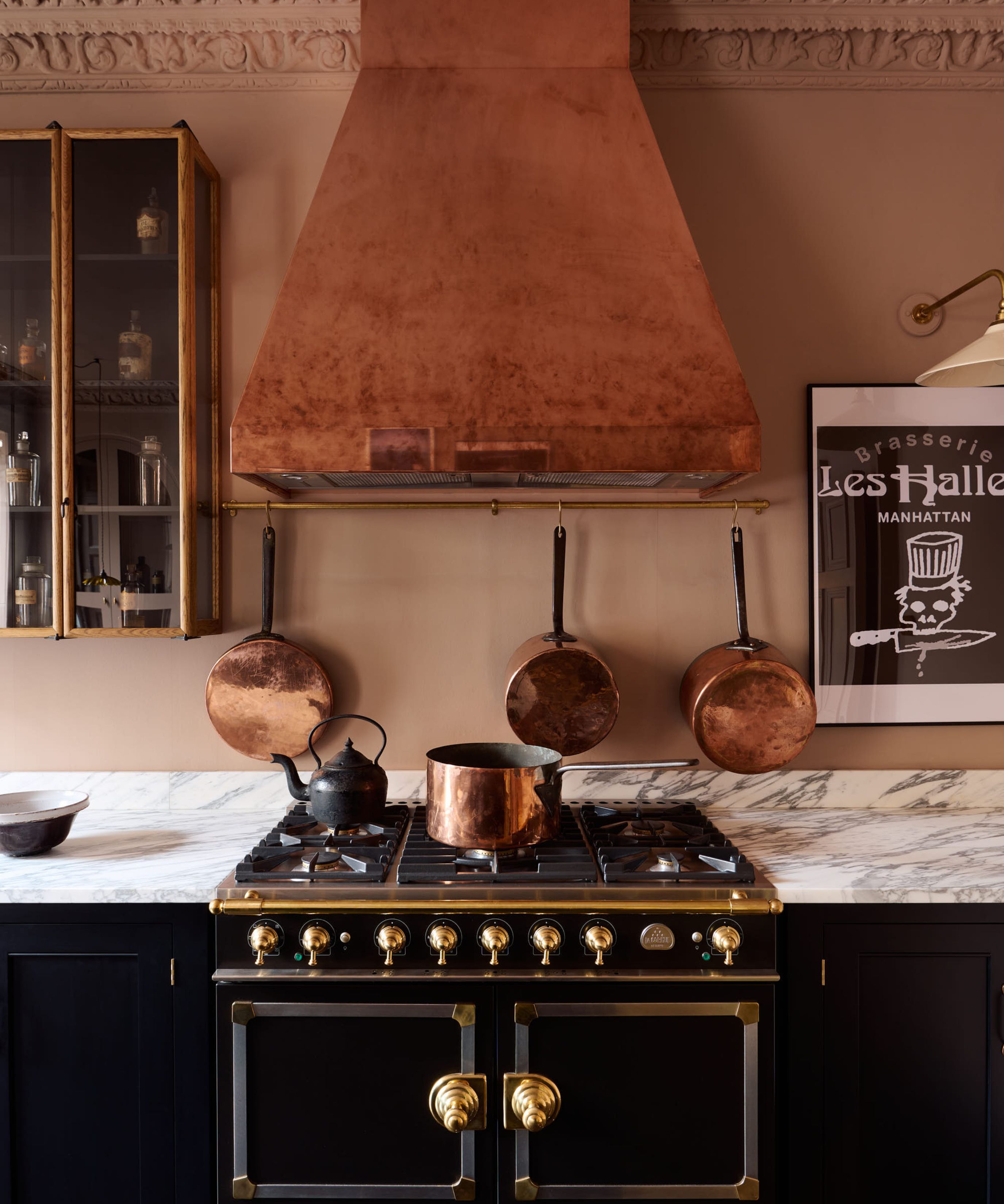 A moody, sophisticated kitchen featuring a large copper range hood over a professional black and gold stove. Copper pots hang from a brass rail against dusty rose walls, complemented by a white marble countertop.